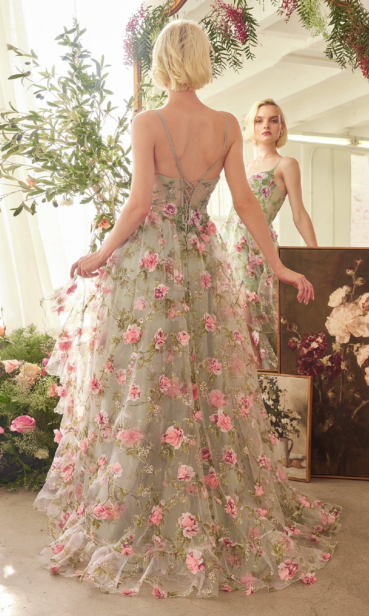 A woman wearing the Sage Long Floral Prom Dress by Andrea & Leo (A1420), featuring sheer floral details and pink flowers, stands facing a mirror in a sunlit room with plants and floral paintings.
